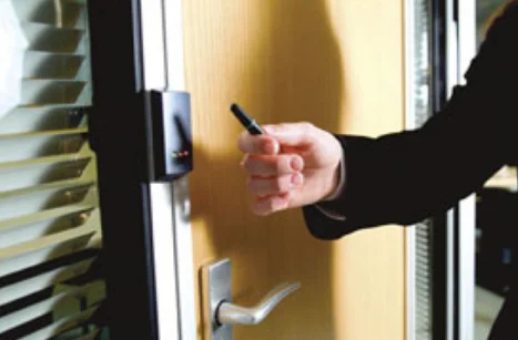 Close-up of a person in a black sleeve using a key card to access a wooden door with a silver handle and electronic access control reader near office blinds.