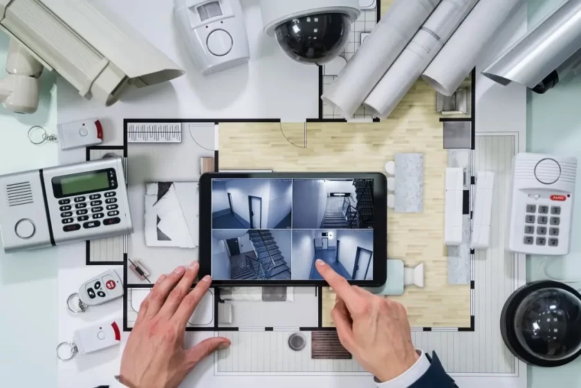 Person viewing security camera footage on a tablet, surrounded by blueprints, security system devices, alarm panels, cameras, and access cards.