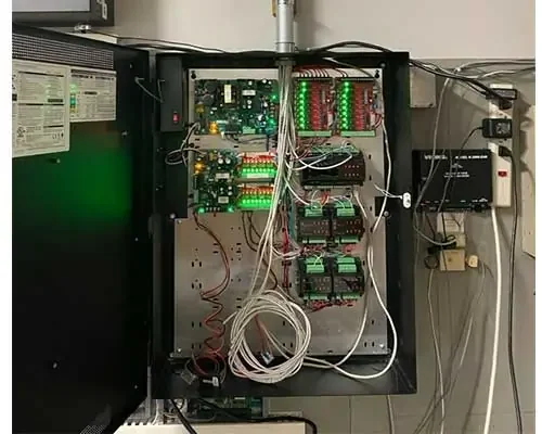 Open electrical control panel with exposed circuit boards, wiring, and indicator lights mounted on a wall.