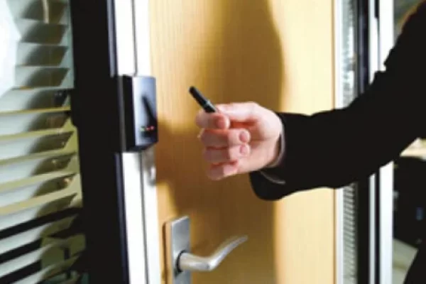 Close-up of a person in a black sleeve using a key card to access a wooden door with a silver handle and electronic access control reader near office blinds.